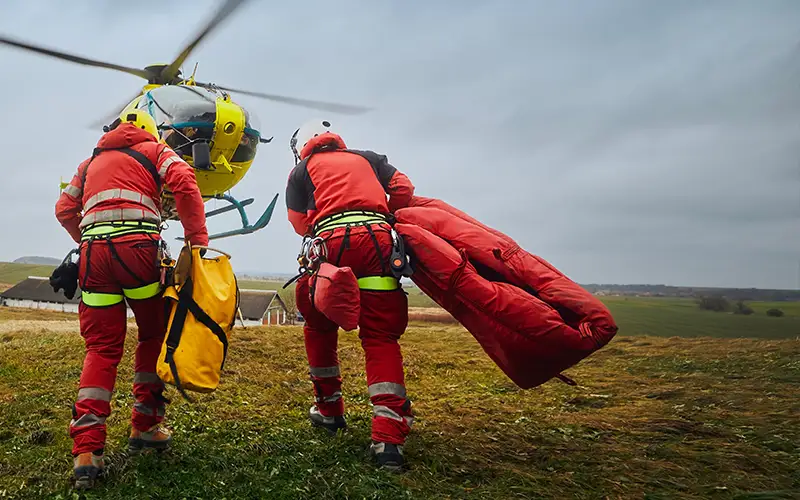 Two men dressed in red and yellow emergency uniforms are lifting a bag