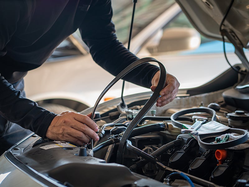 A man repairs a car belt, focused on his work with tools in hand and the hood raised.