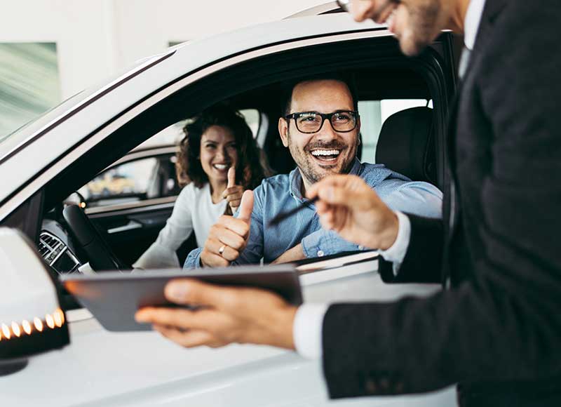 A man and woman in a car smile while the salesperson holding up a tablet, showcasing their excitement.
